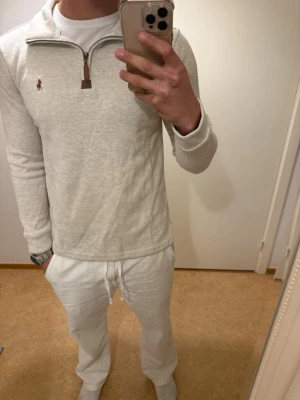 Ljusgrå långärmad polotopp med half zip - Säljer en stilren ljusgrå långärmad polotopp med half zip och liten broderad logga på bröstet. Toppen har en mjuk ribbad struktur och hög krage som kan bäras öppen eller stängd. Perfekt för dig som gillar en clean och enkel look.