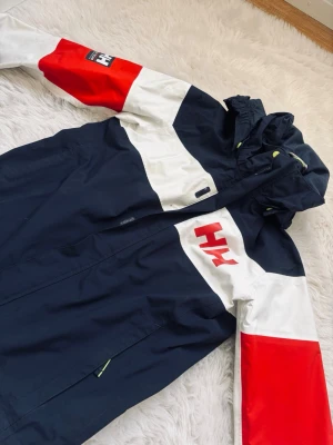 helly hansen vindjacka marin/röd/vit - Snygg vindjacka från Helly Hansen i marinblått, vitt och rött. Jackan har huva, dragkedja framtill och stora HH-loggor på bröstet och ärmen. Färgblockad design och praktiska fickor med dragkedja. Perfekt för en sportig och trendig look.