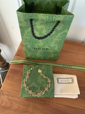 Gucci guldarmband med rosa pärlor - Snyggt armband från Gucci i guldfärgad metall med klassiska GG-loggor och dekorerat med små rosa pärlor. Armbandet har en elegant kedja och detaljer som ger en lyxig känsla. Perfekt accessoar för att lyfta din stil.