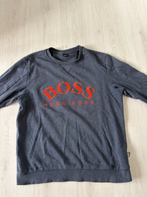 Grå sweatshirt från Hugo Boss - Snygg grå sweatshirt från Hugo Boss med stor röd logga på bröstet. Tröjan har rund halsringning, långa ärmar och ribbade muddar vid ärmslut och nederkant. Perfekt för en avslappnad och trendig look.