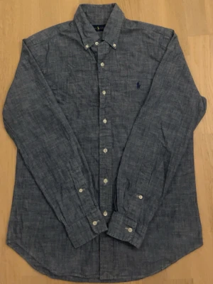 Blå långärmad ralph lauren skjorta - Snygg blå ralph lauren skjorta med klassisk krage och vita knappar. Skjortan har lång ärm och ett diskret broderat märke på bröstet. Perfekt för en stilren look och passar till många olika outfits.