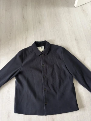 Mörkgrå overshirt från Primark - Stilren mörkgrå overshirt från Primark Edit i regular fit. Skjortjackan har en klassisk krage, knäppning framtill och är tillverkad i ett ribbat tyg som ger en modern känsla. Perfekt att slänga över en t-shirt för en avslappnad look.