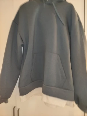 Grå hoodie med magficka och huva - Säljer en stilren grå hoodie med klassisk magficka och huva med dragsko. Hoodien har ribbade muddar vid ärmslut och nederkant samt en vit detalj längst ner. Perfekt för en avslappnad och trendig look.