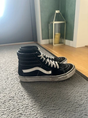 Svarta höga sneakers från Vans - Klassiska svarta höga sneakers från Vans med vit sula och vit sidostrip. Skorna har vit snörning och kontrastsömmar. Ovandelen är i mocka och textil, vilket ger en skön och stilren look. Perfekta för dig som gillar streetwear och skatekänsla.