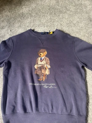Mörkblå sweatshirt Polo Bear Ralph Lauren - Säljer en mörkblå sweatshirt från Ralph Lauren med det ikoniska Polo Bear-trycket på bröstet. Tröjan har rund halsringning och ribbade muddar vid ärmslut och nederkant. Tröjan är inprincip helt oanvänd och därför i nyskick.
