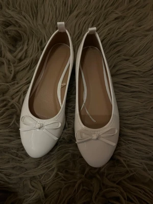 Vita ballerinaskor med rosett - Säljer ett par klassiska vita ballerinaskor i glansigt material med rund tå och söt rosettdetalj framtill. Skorna har en enkel och stilren design med små öglor bak för enkel påtagning. Perfekta för dig som gillar en clean och feminin look.