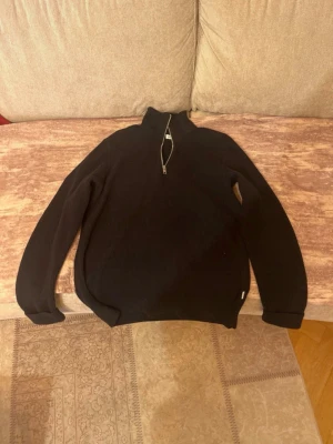 Svart stickad tröja med halv dragkedja - Svart stickad tröja med hög krage och halv dragkedja framtill. Tröjan har långa ärmar och ribbade muddar vid ärmslut och nederkant. Perfekt för kyliga dagar och enkel att matcha med olika outfits.