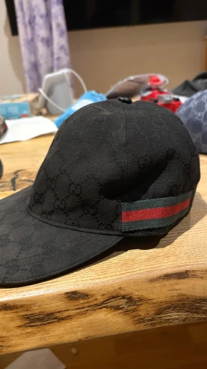 Gucci keps - Snygg svart keps från Gucci med det klassiska GG-monogrammönstret över hela kepsen. På sidan finns en grön och röd rand som signaturdetalj. Justerbar rem baktill för perfekt passform. Tillverkad i slitstarkt tyg som känns lyxigt.