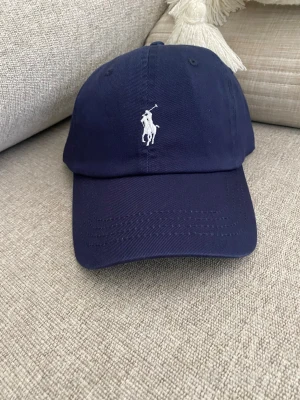 Mörkblå keps från Polo Ralph Lauren unisex - Snygg mörkblå keps från Polo Ralph Lauren med klassisk vit broderad logga framtill och 'POLO' broderat bak. Justerbar rem baktill för perfekt passform. Tillverkad i bomullstyg som andas och passar till de flesta outfits.