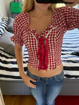 Rutig röd blus med snörning - Supersöt rutig blus i rött och vitt med snörning framtill och puffiga korta ärmar. Toppen har en croppad passform och dragkedja i sidan. Perfekt för dig som gillar vintagevibbar och detaljer som sticker ut.