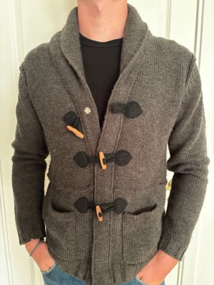 Grå cardigan  - Mysig kofta grovstickad känsla och stora träknappar i toggle-stil. Koftan har två framfickor, bred sjalkrage och svarta detaljer vid knäppningen. Perfekt för lager på lager och en avslappnad look.