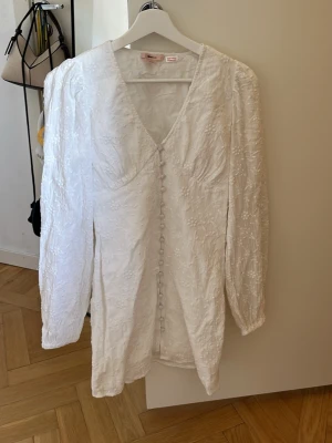 Vit kortklänning från Gina Tricot - Supersöt vit kortklänning från Gina Tricot med v-ringning, långa ärmar och knappar framtill. Klänningen har ett diskret blommigt mönster i tyget och lätt puffade axlar. Perfekt för sommarens alla tillfällen! Använd 1 gång