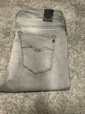 Grå slim fit jeans från Replay hyperflex - Snygga grå slim fit jeans från Replay med klassisk femficksmodell och diskret slitning. Jeansen har en stilren bakficka med broderad detalj och Replay-logga på linningen. Perfekta för en trendig och avslappnad look. Passar både kille och tjej 