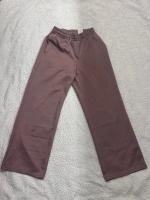 mjukisbyxor från H&M - Säljer mjukisbyxor från H&M med bred resår i midjan och dragsko. Byxorna har raka ben och är gjorda i ett mjukt, slätt material som känns skönt mot huden. Perfekta för chill dagar eller när du vill vara extra bekväm. Gammelrosa-brun