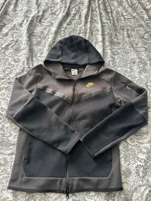 Mörkgrå Nike hoodie med dragkedja - Säljer en mörkgrå hoodie från Nike med dragkedja framtill och gul logga på bröstet. Hoodien har huva, två fickor och diskreta reflexdetaljer över bröstet. Tillverkad i mjukt bomullsmaterial som är skönt att ha på sig.