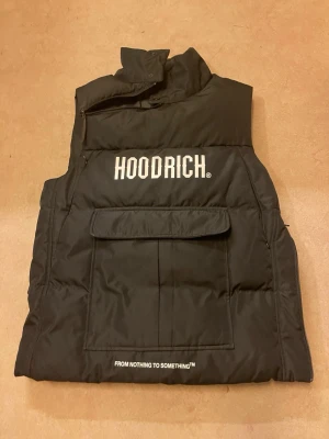 Svart dunväst från Hoodrich - Snygg svart dunväst från Hoodrich med stor ficka framtill och vit logga på bröstet. Västen har hög krage och dragkedja i sidan för extra stil och funktion. Materialet är vadderat och ger en modern streetwear-look.