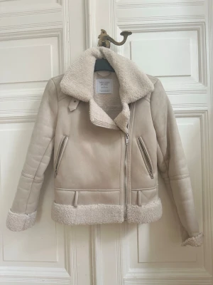 Beige teddyjacka med dragkedja - Snygg beige teddyjacka med stor krage och detaljer i fuskpäls. Jackan har två snedställda fickor med dragkedja, breda muddar och markerad nederkant i teddy. Perfekt för kalla dagar och ger en trendig look.