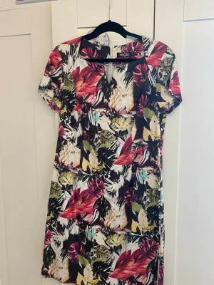 Färgglad blommig kortklänning - Klänning med tropiskt blommönster i rött, grönt, svart, vitt och inslag av gult och blått. Klänningen har en liten v- ringning och korta ärmar. Den har också en dragkedja på baksidan. Perfekt för sommarens varma dagar och ger en fräsch look.                                                      Är öppen för att förhandla priset🌸
