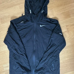 Svart vindjacka från Nike med huva - Snygg svart vindjacka från Nike med huva och dragkedja framtill. Jackan har två sidofickor med dragkedja och reflekterande Nike-logga på bröstet. Perfekt för blåsiga dagar och sportiga outfits.