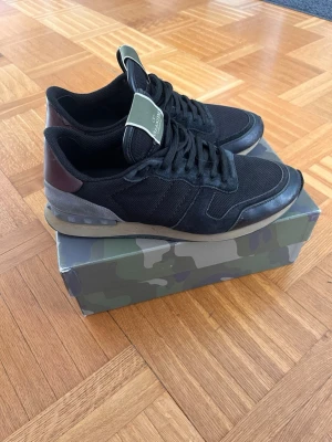 Svarta Valentino sneakers - Snygga svarta sneakers från Valentino med detaljer i mocka och mesh. Box ingår! Skorna har en mörkgrön plös med logga, svarta skosnören och en beige sula. Baktill finns en brun detalj i läder. Perfekta för dig som vill ha en stilren och trendig look.