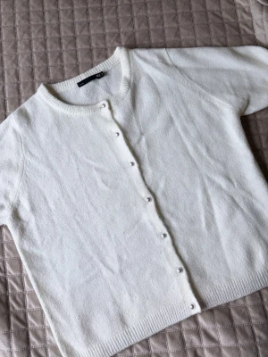 Vit basic kofta med rund hals - En stilren vit kofta med rund halsringning och knäppning framtill. Koftan har långa ärmar och ribbade muddar vid ärmslut och nederkant. Perfekt att slänga över en t-shirt eller klänning för en clean look.