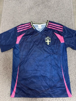 Blå fotbollströja med rosa detaljer - Snygg blå fotbollströja med rosa ränder och detaljer på axlar och sidor. Tröjan har V-ringning med vit och svart kant samt ett klubbmärke med svensk flagga på bröstet. Tillverkad i ett lätt och ventilerande material, perfekt för träning eller match.