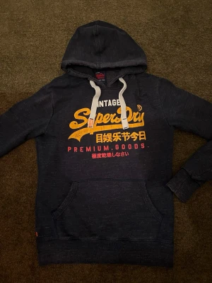 Mörkblå Superdry hoodie med tryck - Snygg mörkblå hoodie från Superdry med stort gult och vitt tryck på bröstet samt röda japanska tecken. Klassisk känguruficka framtill, vita dragsnören i huvan och mjukt material. Perfekt för en avslappnad streetwear-look.