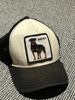 Svart och vit keps med hundmotiv - Cool keps i svart och vitt med broderad patch framtill som visar en rottweiler och texten 'BAD BOY'. Kepsen har nät på sidorna och justerbar snapback. Märkt med 'THE FARM' på insidan. Perfekt för dig som gillar streetstyle och djurmotiv.