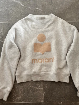 Grå sweatshirt från Isabel Marant - Säljer en ljusgrå sweatshirt från Isabel Marant Étoile i 100% bomull. Tröjan har en beige logga och text framtill, ribbade muddar och en avslappnad  passform. Perfekt för en chill och trendig look. Köpt på Vinted så kan tyvärr inte garantera äkthet men ser äkta ut! Har fått lite smink på sig men går säkert att få bort! Pris kan diskuteras 💕 Säljer många populära och eftertraktade plagg så kolla in min profil!