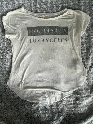 Vit Hollister t-shirt med tryck - Vit t-shirt från Hollister med trycket 'Hollister Los Angeles' i grått framtill. T-shirten har rund halsringning och korta ärmar. Tillverkad i mjuk bomull, perfekt för en avslappnad och enkel stil.