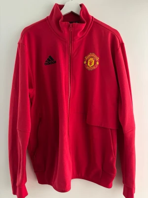 Röd Manchester United ziptröja Adidas - Snygg röd ziptröja från Adidas med Manchester Uniteds klubbmärke på bröstet och Adidas-logga i svart. Tröjan har hög krage, hel dragkedja och ribbade muddar. Perfekt för träning eller att bära till vardags. Tillverkad i mjukt material.
