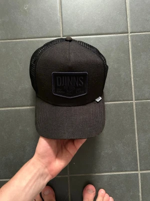 Svart truckerkeps från DJINNS - Säljer en svart truckerkeps från DJINNS med mesh på sidorna och justerbar snapback. Kepsen har en broderad patch framtill och är tillverkad i en mix av bomull och polyester. Perfekt för en avslappnad streetwear-look.