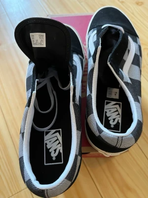 Vans slip-on svartvitrutiga sneakers - Säljer ett par klassiska Vans slip-on sneakers i svart och vitt rutigt mönster. Skorna har vit sula, svart tåparti och är tillverkade i canvasmaterial. Perfekta för dig som gillar streetwear och vill ha en tidlös look.
