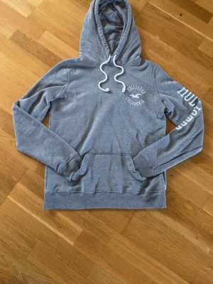 Grå hoodie från Hollister - Snygg grå hoodie från Hollister med vit logga på bröstet och text på ena ärmen. Klassisk känguruficka framtill, ribbade muddar och justerbar huva med vita snören. Perfekt för en avslappnad och trendig look. Storlek S