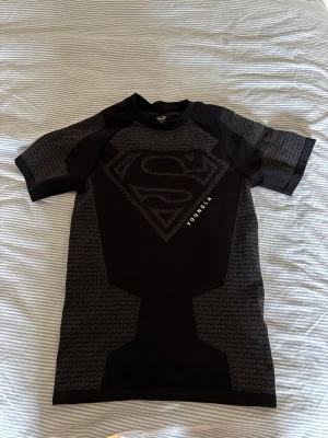 Svart Superman t-shirt från YoungLA - Säljer en svart t-shirt från YoungLA med stort Superman-tryck på bröstet. T-shirten har korta ärmar och coola mönstrade detaljer i grått på ärmar och sidor. Perfekt för dig som gillar superhjältar och vill sticka ut.
