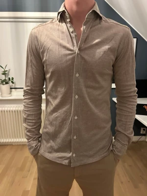 Beige långärmad skjorta från Eton - Säljer en stilren beige skjorta från Eton med klassisk krage och vita knappar. Skjortan är långärmad och har en slim passform, perfekt för dig som gillar en clean och enkel look. Tillverkad i mjuk bomull för skön känsla.