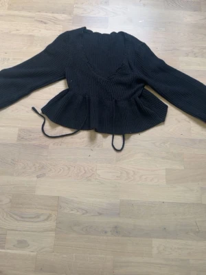Svart peplum stickad tröja - Säljer en svart stickad tröja med peplum och långa ärmar. Tröjan har v-ringning och dekorativ snörning i midjan. Ribbstickad struktur och volang nedtill ger en trendig siluett. Perfekt för dig som vill ha en snygg och bekväm look.