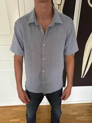 Ljusblå kortärmad skjorta med knappar - Snygg ljusblå skjorta med korta ärmar och klassisk krage. Skjortan har diskret mönster och stängs med ljusa knappar framtill. Perfekt till jeans eller shorts för en avslappnad stil.