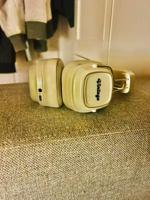Beige over-ear hörlurar Vintage - Stilrena over-ear hörlurar i beige med fyrkantiga kåpor och vadderad bygel. Hörlurarna har texten 'Vintage' på sidan och detaljer i metall. De har en retroinspirerad design och är tillverkade i konstläder och plast.