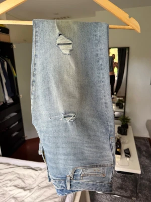 Ljusblå slitna jeans med hål - Säljer ett par ljusblå jeans med trendiga slitningar och hål på benen. Jeansen har klassisk femficksdesign och raka ben. Perfekta för en avslappnad och cool stil. Materialet är mjukt denim i bomull.