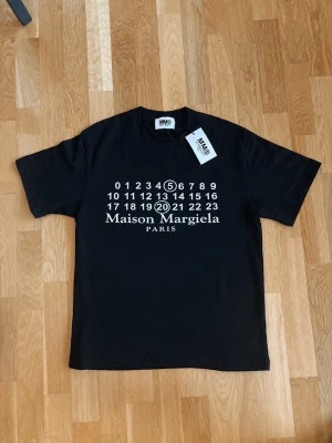 Svart Maison Margiela t-shirt - Snygg svart t-shirt från Maison Margiela med ikoniskt tryck framtill i vitt. Klassisk rund hals och korta ärmar. Perfekt för dig som gillar stilrena och trendiga plagg med en twist. Materialet känns mjukt och skönt mot huden.