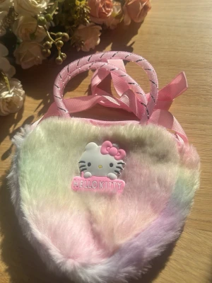 Pastellfärgad Hello Kitty handväska - Supersöt handväska i mjuk pastellfärgad fuskpäls med Hello Kitty-motiv framtill. Väskan har två rosa handtag med glittriga detaljer och en rosa rosett. Perfekt för dig som älskar gulliga accessoarer och vill sticka ut lite extra.