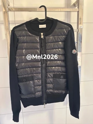 Svart cardiganjacka från Moncler - Snygg svart cardiganjacka från Moncler med quiltad front och två fickor med knappstängning. Ärmar och rygg i mjukt tyg, rund halsringning och dragkedja framtill. Moncler-logga på ena ärmen ger en exklusiv touch.