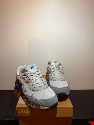 Vita och grå Asics sneakers - Säljer ett par fräscha sneakers från Asics i vitt och ljusgrått med mesh och mockadetaljer. Skorna har klassisk låg siluett, vita snören och blå logga på plösen. Perfekta för dig som gillar stilrena och sportiga vibes.