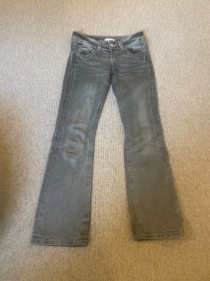 Grå bootcut jeans från Gina Tricot - Säljer ett par grå bootcut jeans från Gina Tricot med snygga slitningar och klassiska fem fickor. Jeansen har vit kontrastsöm och knappgylf. Perfekta för dig som gillar en avslappnad men trendig look.