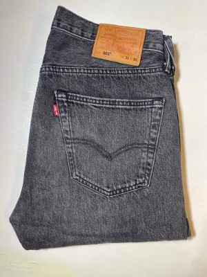 Levi's 501 raka jeans - Säljer ett par klassiska grå Levi's 501 jeans med rak passform och fem fickor. Jeansen har den ikoniska röda Levi's-lappen på bakfickan och patch i läder bak i midjan. Tillverkade i slitstark denim, perfekta för en avslappnad och tidlös stil.