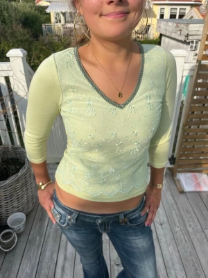 Ljusgrön y2k tröja med spetsdetaljer - Snygg ljusgrön blus med lång ärm och v-ringning. Framsidan har ett tunt lager spets med broderade blommor och vågig kant nedtill. Perfekt för dig som gillar 2000s och mjuka färger.