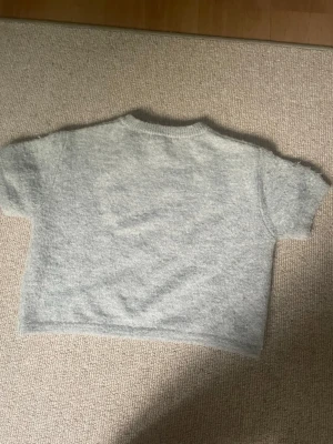 Grå kortärmad stickad tröja - Mysig grå stickad tröja med korta ärmar och rund hals. Tröjan har en boxig, croppad passform och är tillverkad i ett mjukt material som känns skönt mot huden. Perfekt att styla över en skjorta eller med höga jeans.
