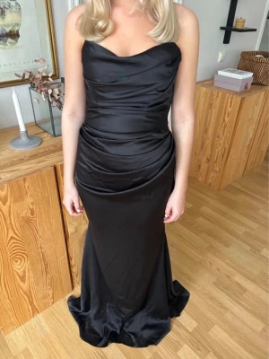 Svart långklänning i satin - Elegant svart långklänning i glansigt satinmaterial med draperad överdel och figurnära passform. Klänningen är axelbandslös och har en smickrande siluett som faller snyggt mot golvet. Perfekt för speciella tillfällen!