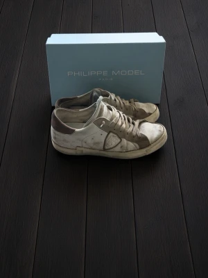 Vita Philippe Model sneakers - Snygga vita sneakers från Philippe Model med beige och bruna detaljer. Klassisk låg modell med vita snören och diskret logga på sidan. Tillverkade i läder med gummisula. Perfekta för en stilren och avslappnad look.